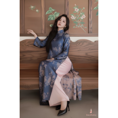 A woman wearing a blue floral vietnamese dress with a pink inner skirt sits on a wooden bench in front of a wooden wall with floral paintings.