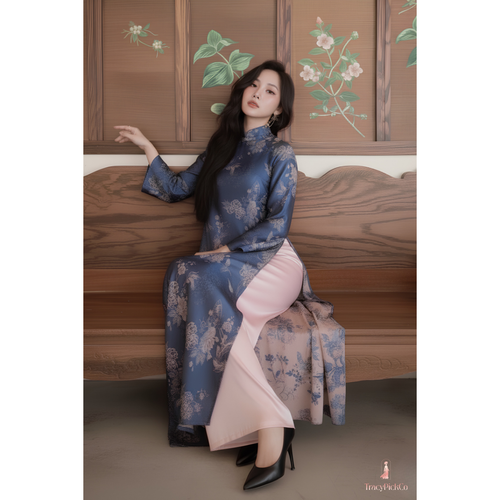 A woman wearing a blue floral vietnamese dress with a pink inner skirt sits on a wooden bench in front of a wooden wall with floral paintings.