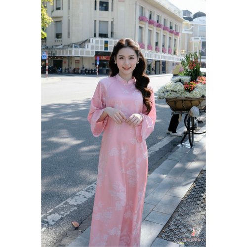 A woman wearing a pink traditional Vietnamese ao dai dress with a subtle floral pattern, standing on a city street with buildings and a flower cart in the background.